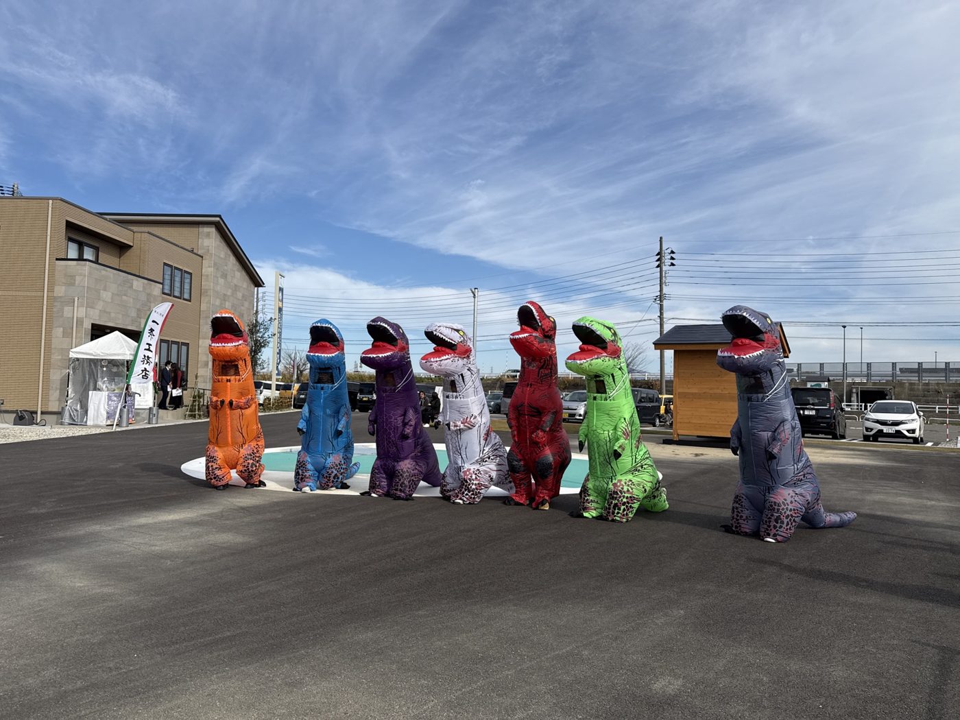 住宅公園の広場で並ぶ、色とりどりの恐竜の着ぐるみ7体（前回のレース）